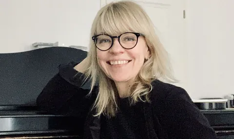 Eine lächelnde Frau mit blonden Haaren und Brille am Klavier.