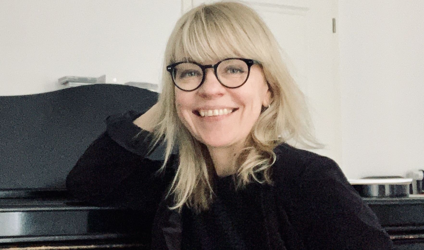 Eine lächelnde Frau mit blonden Haaren und Brille am Klavier.